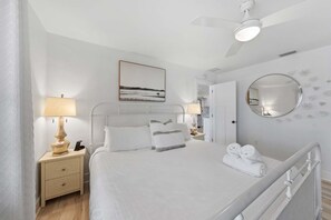 A hotel room with a bed, bedside tables, a ceiling fan, a painting, a mirror, and a view of the hallway. - Serendipity Gulfront - Hot Tub - Heated Pool -golf Cart (Anna Maria)