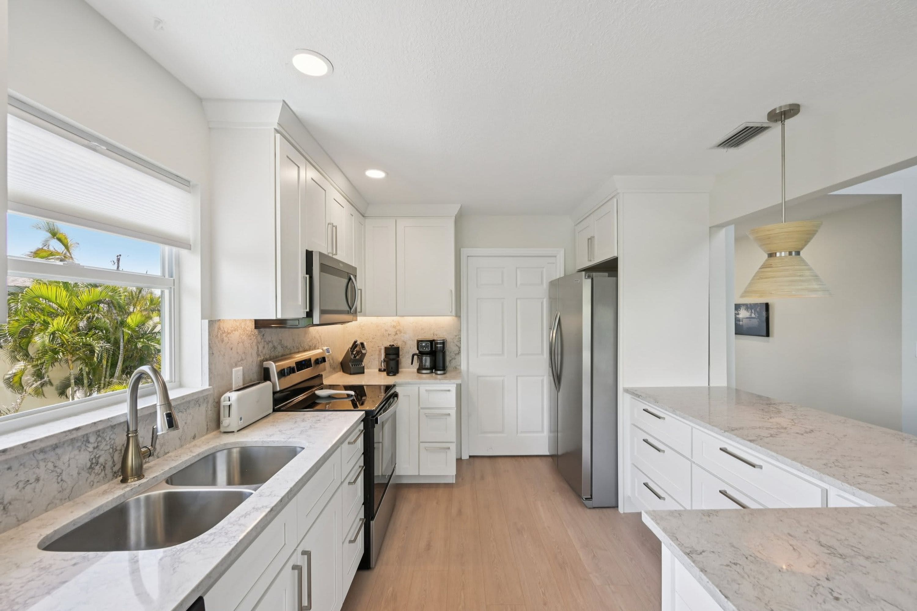 A modern kitchen with white cabinetry, stainless steel appliances, a marble countertop, and a view of palm trees outside.