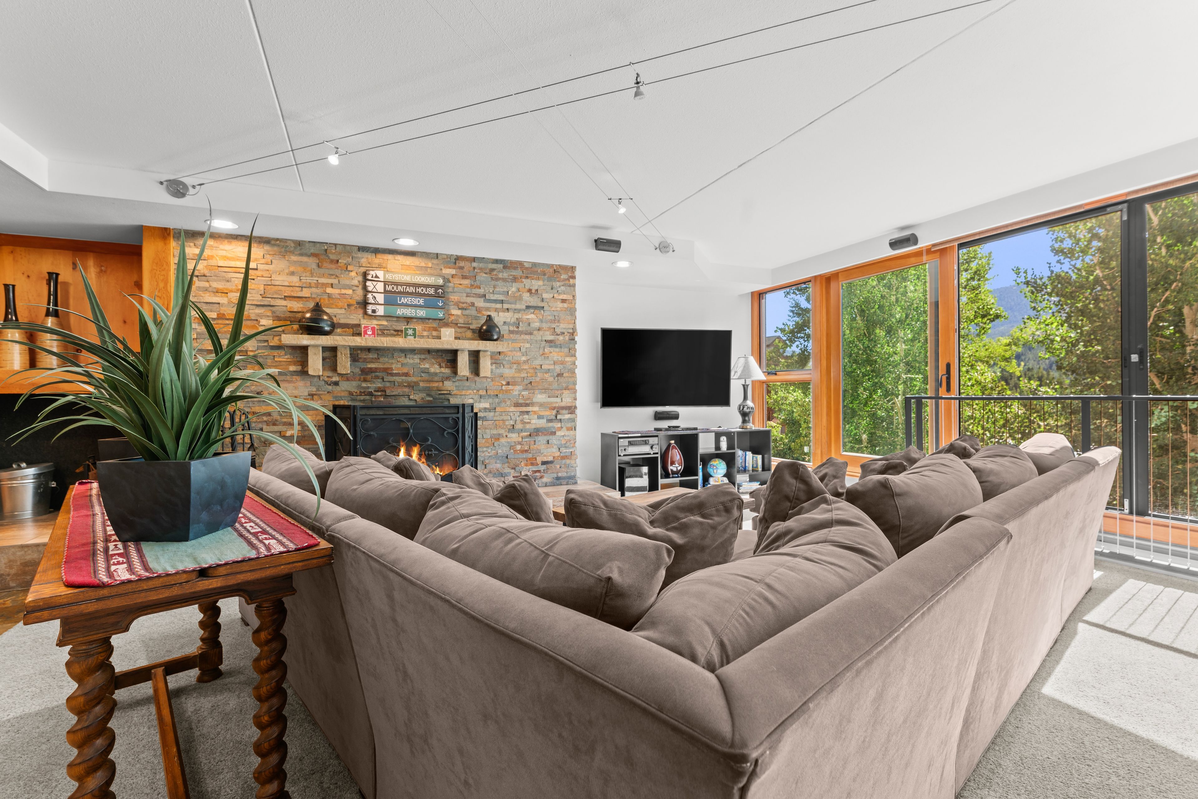 A modern living room with a large sectional sofa, a fireplace, a flat-screen TV, and a balcony with a view of trees and mountains.