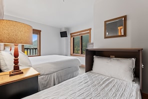 A hotel room with two beds, a bedside table with a lamp, a TV mounted on the wall, and a window with a view of trees. - Rh2284-1 (Keystone)
