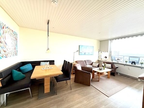 A modern living room with a wooden dining table, black sofas, and a large window offering a view. - Uthlande Holiday Apartment 14 (Wyk auf Foehr)