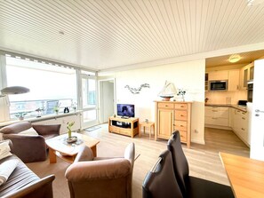 A modern living room with a sofa, coffee table, and TV, a small kitchen with cabinets and appliances, and a balcony with a view. - Uthlande Holiday Apartment 14 (Wyk auf Foehr)
