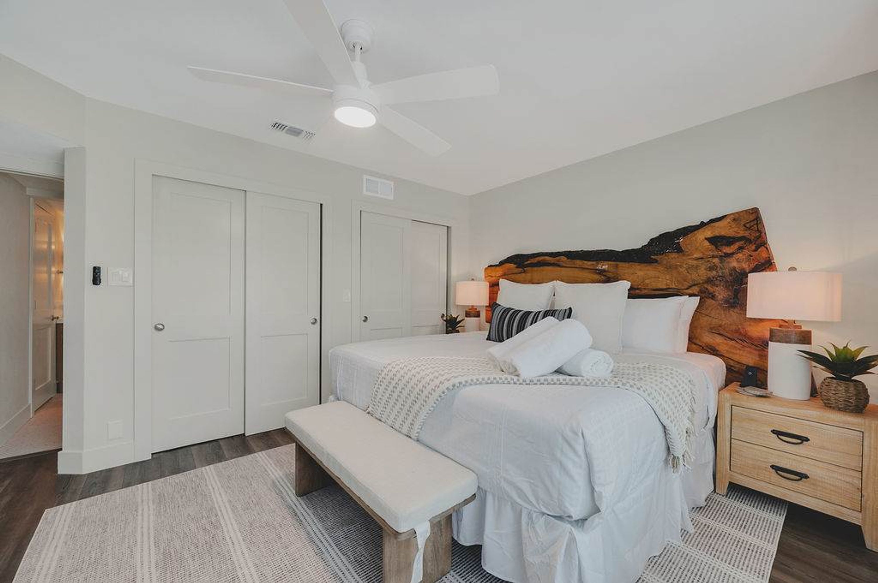 A bedroom with two beds, a ceiling fan, a bench, a nightstand with a lamp, and a large abstract painting on the wall.