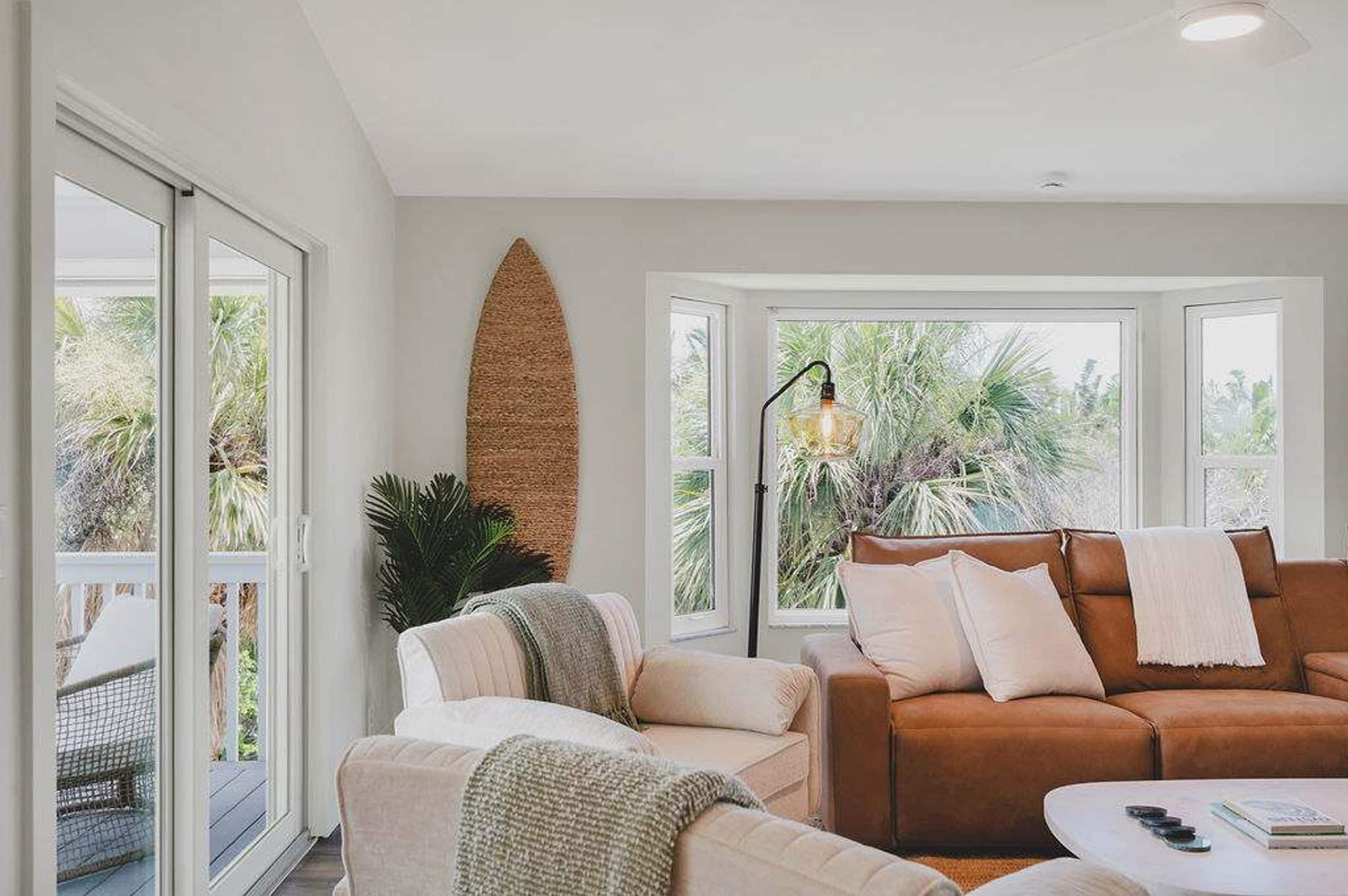 A living room with a brown leather sofa, a white armchair, and a white coffee table. There is a large window with a view of palm trees and a ceiling light fixture.