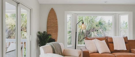 Una sala de estar con un sofá de cuero marrón, un sillón blanco y una mesa de centro blanca. Hay un ventanal con vistas a palmeras y un lámpara en el techo.
