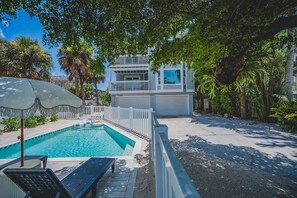 A swimming pool with a lounge chair and umbrella, surrounded by a white fence and lush greenery. - Sandcastle -steps to Beach -pool - Golf Cart (Anna Maria)