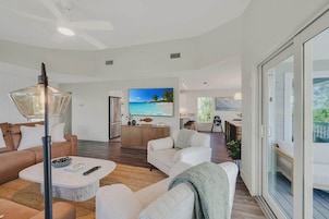 A spacious living area with a large flat-screen TV, a ceiling fan, and a view of the ocean through a sliding glass door.