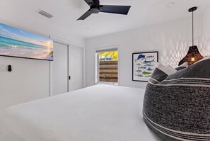 A bedroom with a large bed, a ceiling fan, a painting of a beach, and a framed fish illustration. - Rod Reel Retreat - Heated Pool Spa - Golf Cart - Steps to Beach (Anna Maria)