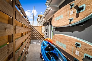 A narrow alleyway with wooden fences on one side and a building with a sloped roof on the other. There are two kayaks with 'KAYAKS' written on them, and a blue bag with a black strap is visible on the ground. - Rod Reel Retreat - Heated Pool Spa - Golf Cart - Steps to Beach (Anna Maria)
