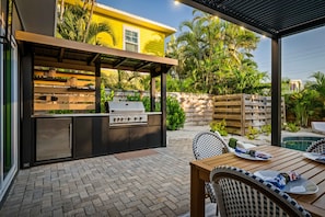 An outdoor kitchen area with a grill, a dining table with chairs, and a pool in the background. - Rod Reel Retreat - Heated Pool Spa - Golf Cart - Steps to Beach (Anna Maria)