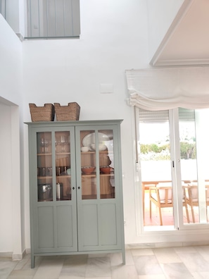 A light green cabinet with glass doors containing kitchenware, a white wall, and a sliding glass door leading to an outdoor patio area. - New Listing (Rota)