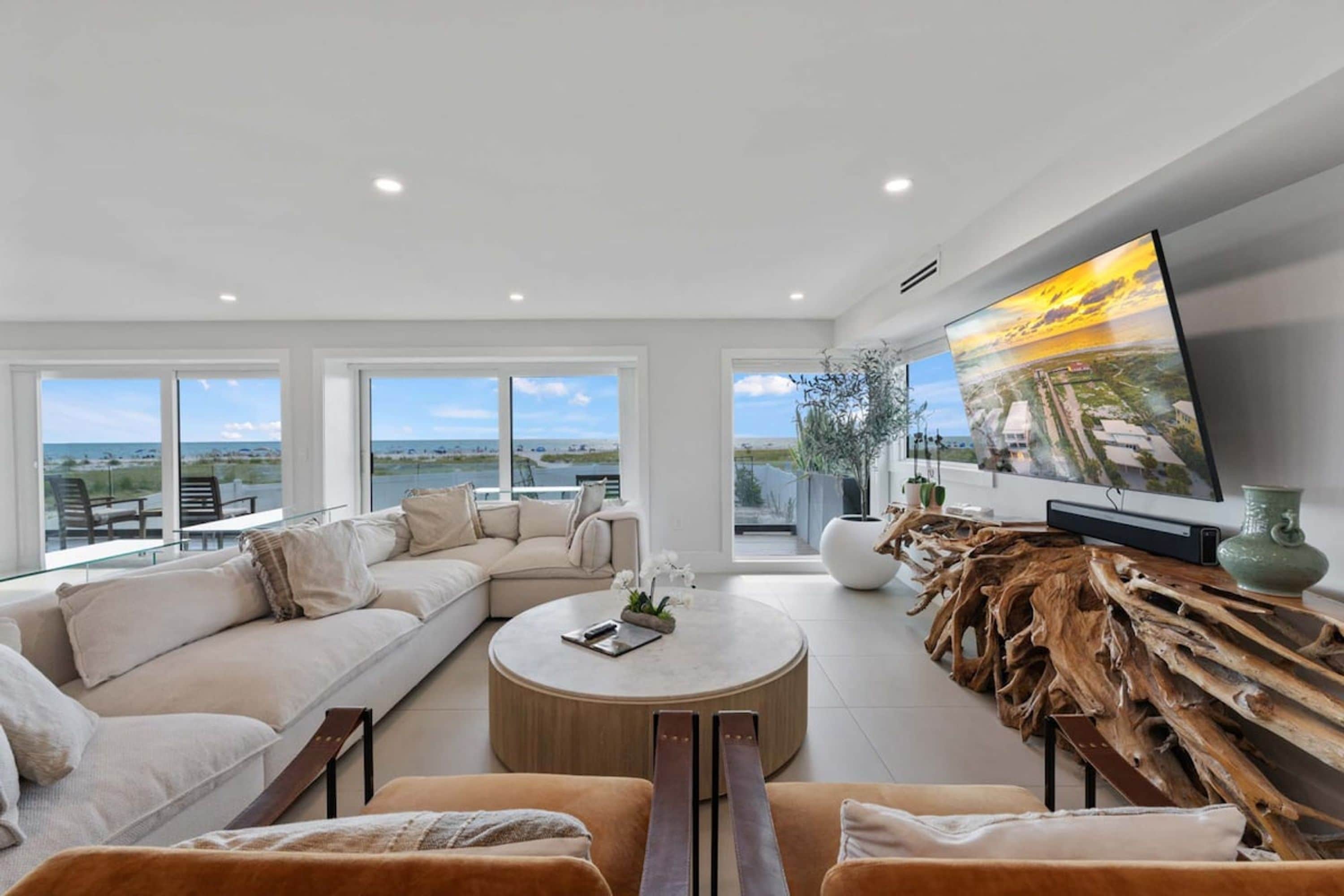 A modern living room with a large flat-screen TV, a beige sectional sofa, a round coffee table, and a view of the ocean through large windows.