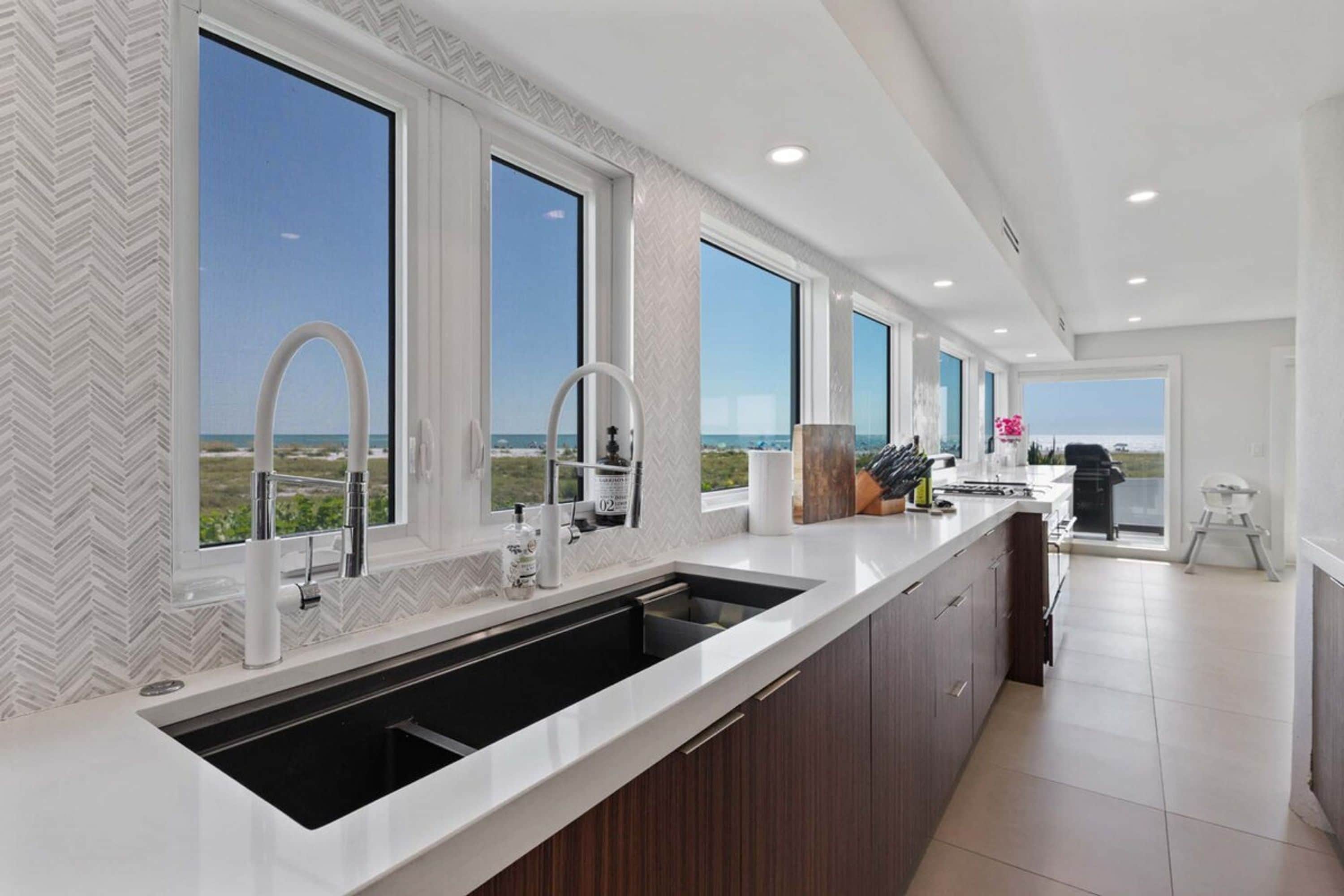 A modern kitchen with a large island, a built-in sink, and large windows offering a view of the beach.