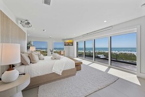 Un dormitorio moderno con una cama grande, una mesita de noche, una lámpara y vistas a la playa a través de amplias ventanas.