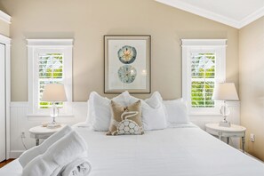 A hotel room with a large bed, two bedside tables, a framed artwork, and windows with a view of greenery. - The Salty Fern - Pool -steps to Beach - Golf Cart (Anna Maria)