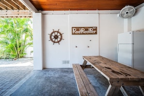 A covered outdoor area with a wooden bench, a wall-mounted sign, a wall-mounted fan, and a view of greenery. - The Salty Fern - Pool -steps to Beach - Golf Cart (Anna Maria)