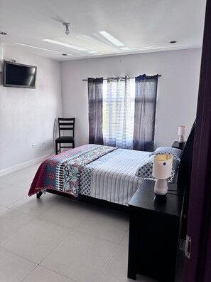 A bedroom with a bed, a chair, a bedside table with a lamp, and a TV mounted on the wall. - Discover Jam Oasis, Tobago! (Mount Pleasant)