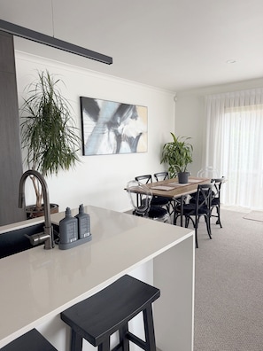 A modern kitchen with a white countertop, a sink, and a dining area with a wooden table and chairs. There are two potted plants, one on the countertop and one on the dining table. - 
The Courtenay - Luxury Urban Retreat (New Plymouth)
