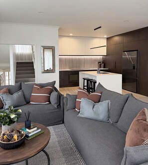 A modern living room with a grey sofa, a coffee table with decorative items, and a view into a kitchen area with a refrigerator and a dining area. - 
The Courtenay - Luxury Urban Retreat (New Plymouth)