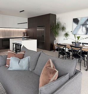 A modern living room with a grey sectional sofa, a dining area with a wooden table and chairs, and a kitchen with dark cabinetry and a built-in fridge. - 
The Courtenay - Luxury Urban Retreat (New Plymouth)
