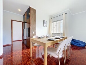 A dining area with a wooden table set for four, a large window with curtains, and a herringbone-patterned floor. - Palmeira Apartment by Atlantic Holiday (Santa Cruz)