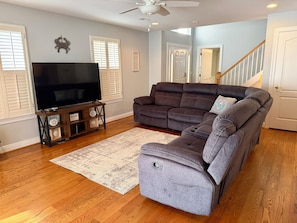 A living room with a large flat-screen TV, a gray sectional sofa, a wooden entertainment unit, and a white rug. - Beautiful 4 bedroom home on the bike trail and east of Rt 1! (Lewes)