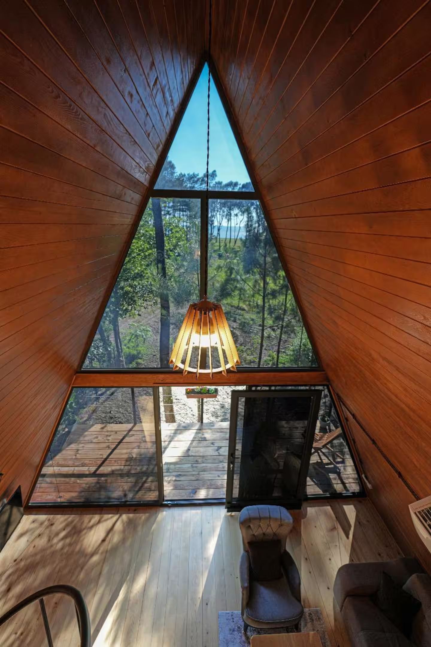 A triangular wooden room with a hanging light fixture and a view of the forest.