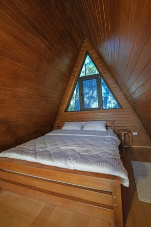 A wooden interior with a sloped ceiling, a bed with white bedding, and a window with a view of trees.