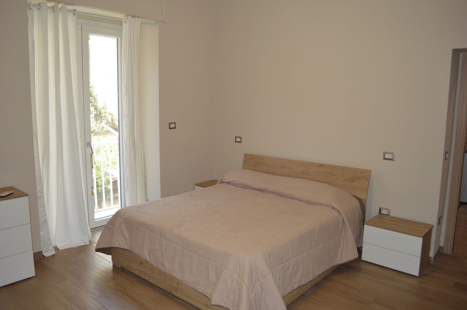 A bedroom with a bed, two bedside tables, a window with curtains, and a door.