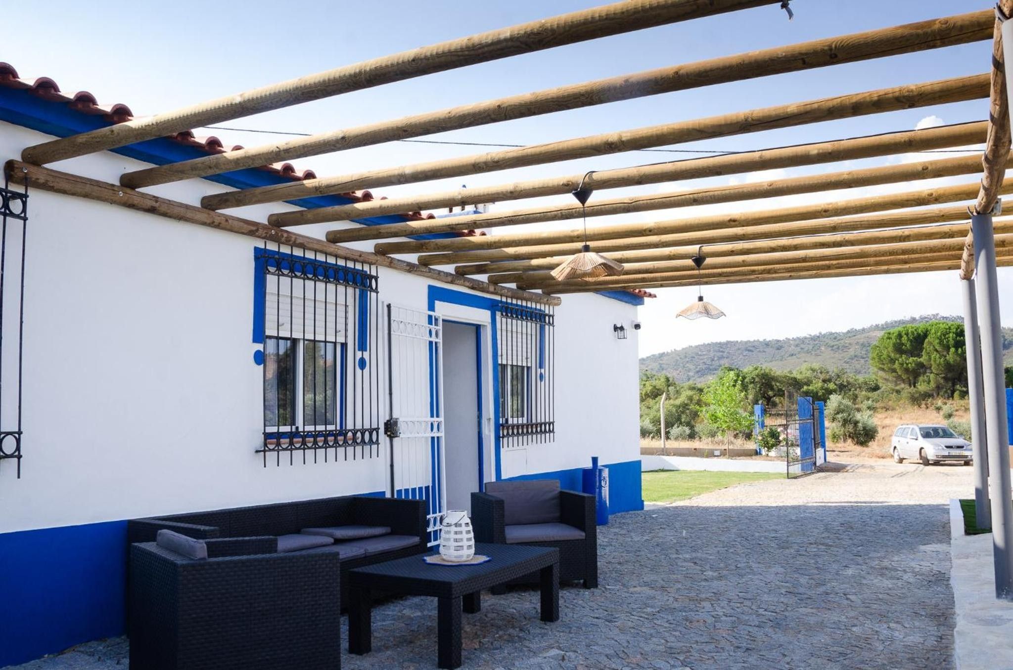 Zona d'estar exterior coberta amb taula i cadires, una pèrgola a sobre i una vista cap a un cotxe i vegetació al fons.