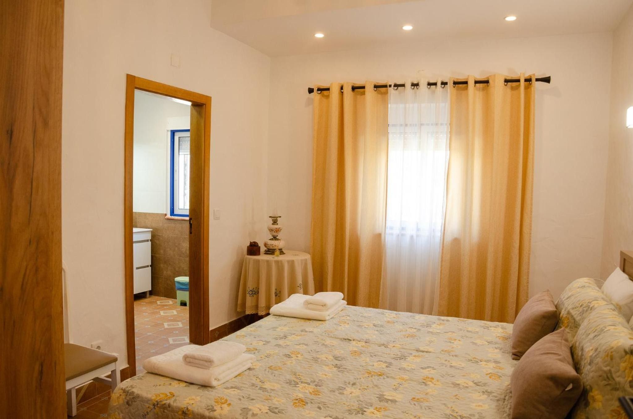 Un dormitori amb un llit, una taula de nit, un mirall, una finestra amb cortines, i un bany visible per una porta oberta.