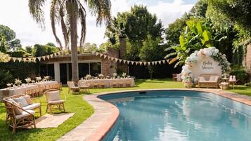An outdoor pool area with lounge chairs and tables, surrounded by greenery and a covered seating area.