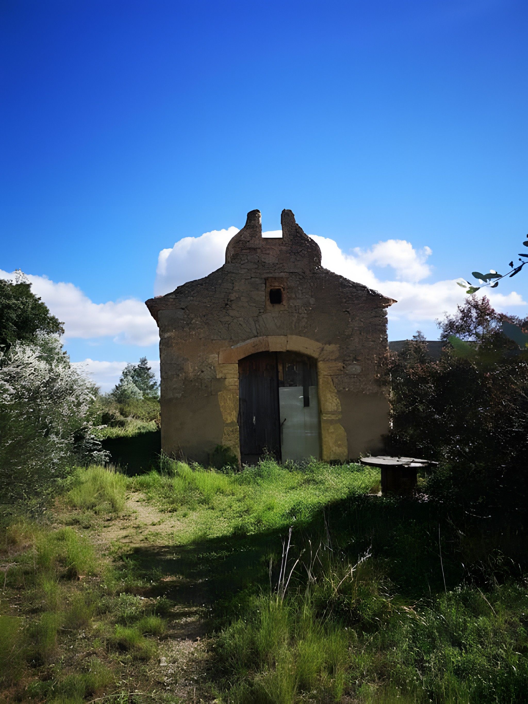 Un pequeño edificio de piedra con una sola puerta y una ventana pequeña, rodeado de follaje y un cielo despejado.