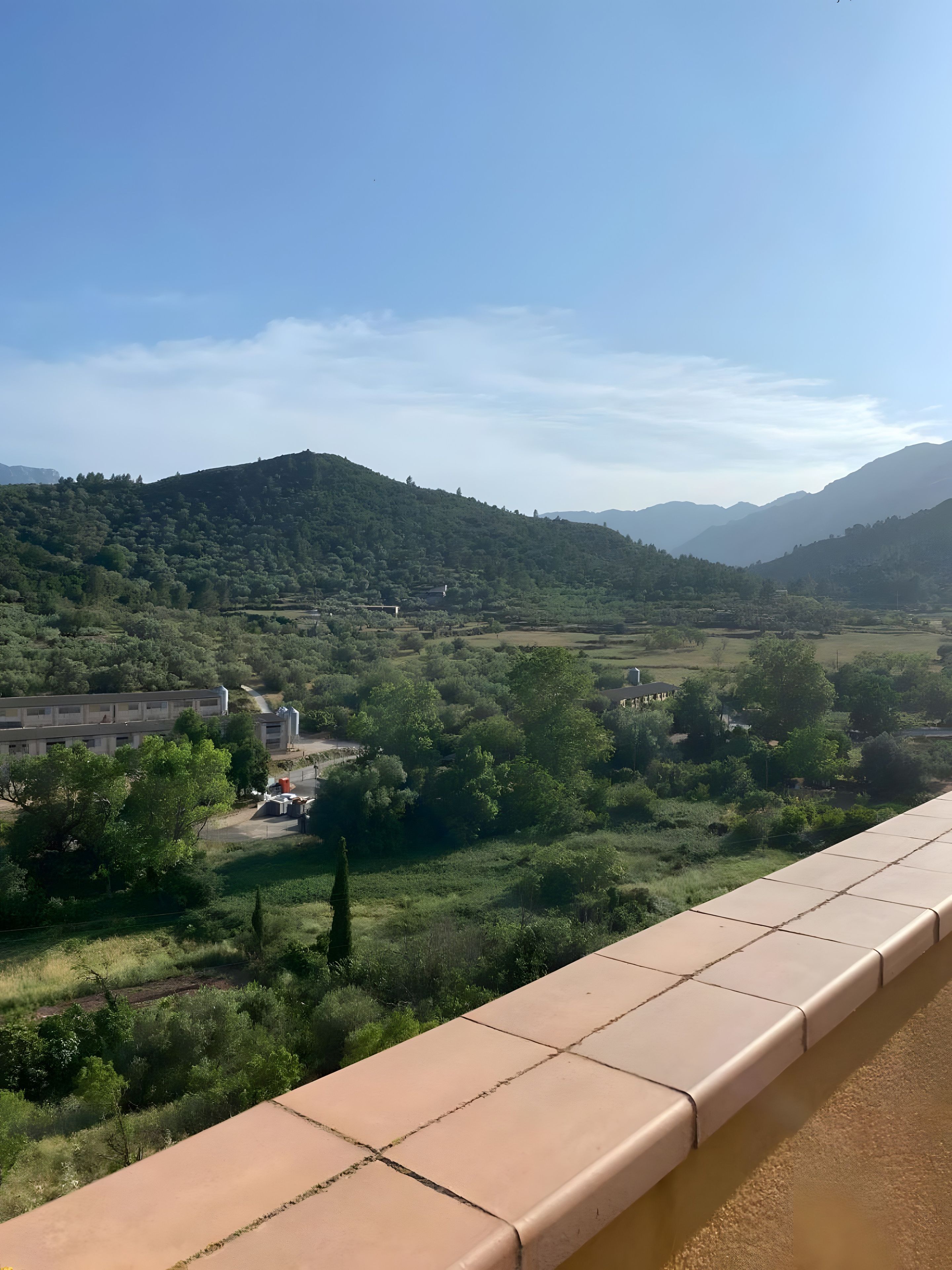 Vistes des d'un balcó sobre una vall amb muntanyes a la distància, un edifici amb un siló i un cel serè.