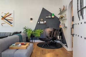 A modern living room with a grey sofa, a black chair, and a wooden coffee table. There are two potted plants, a black rug, and a wall-mounted shelf with books and decorative items. - Hauser House l Artful Spanish-Style Bungalow (Los Angeles)