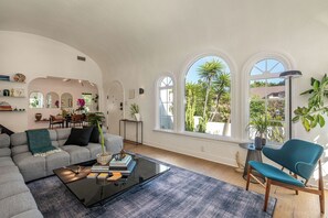 A spacious living room with a grey sofa, a coffee table, and a blue armchair. There are large windows with views of greenery outside. - Hauser House l Artful Spanish-Style Bungalow (Los Angeles)
