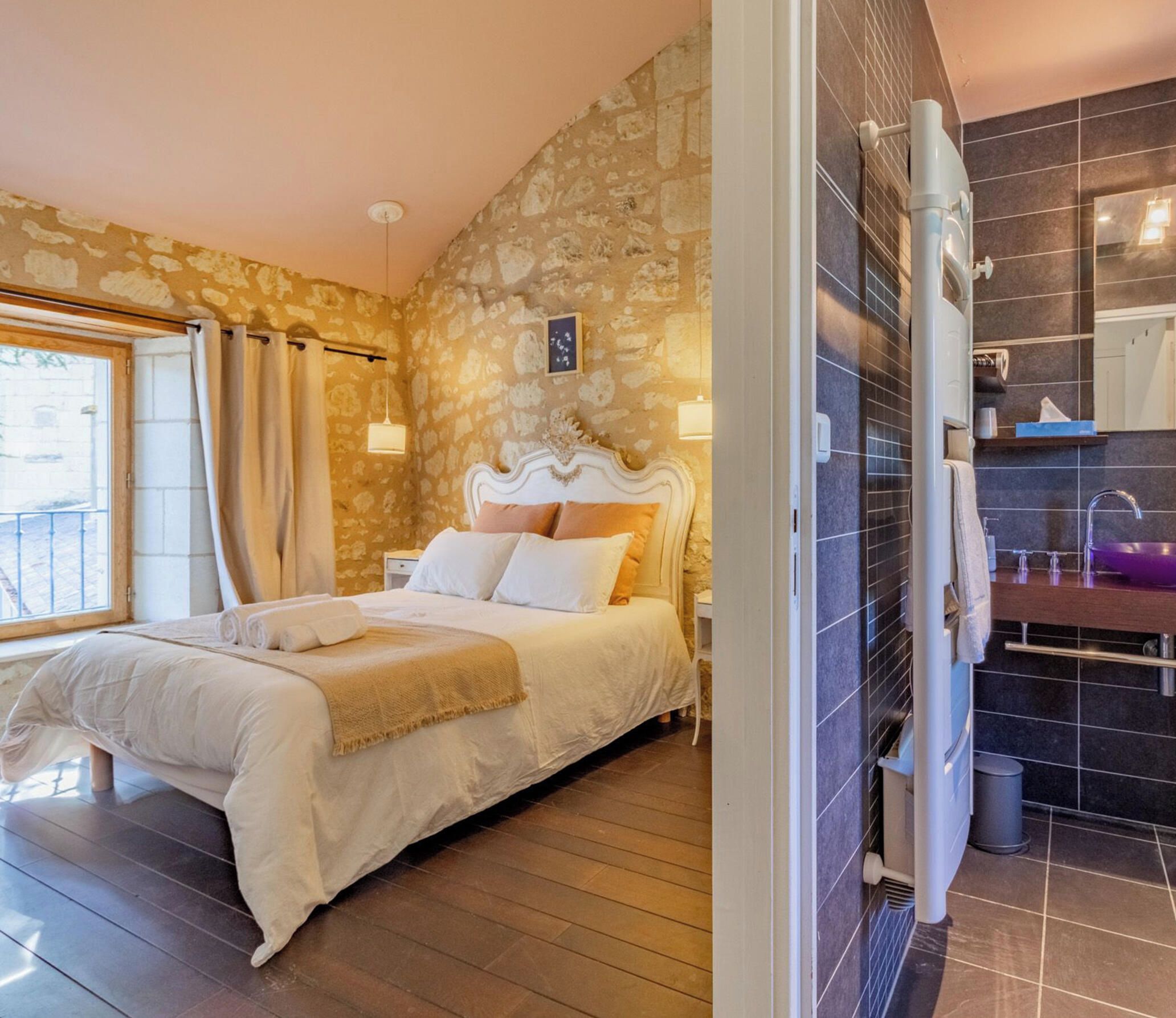 A bedroom with a stone wall, a bed with white bedding, a wooden floor, and a bathroom with dark tiles.