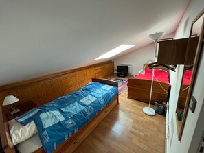 A small, single-room apartment with a sloped ceiling, a bed with a blue patterned bedspread, a desk with a lamp, a television, and a small table with a colorful container. - Family duplex, pets welcome, parking - 6 guests (Brides-les-Bains)