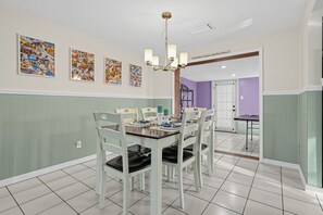 Un comedor con una mesa dispuesta para cuatro personas, rodeada de sillas blancas con cojines negros. Hay tres fotografías enmarcadas en la pared sobre la mesa.