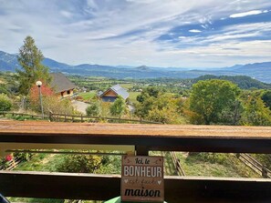 A scenic view from a balcony with a wooden railing, overlooking a valley with houses and mountains. A sign in the foreground reads 'ici, le BONHEUR est fait maison.' - Cosy studio with south balcony - 2 guests (Saint-Michel-de-Chaillol)