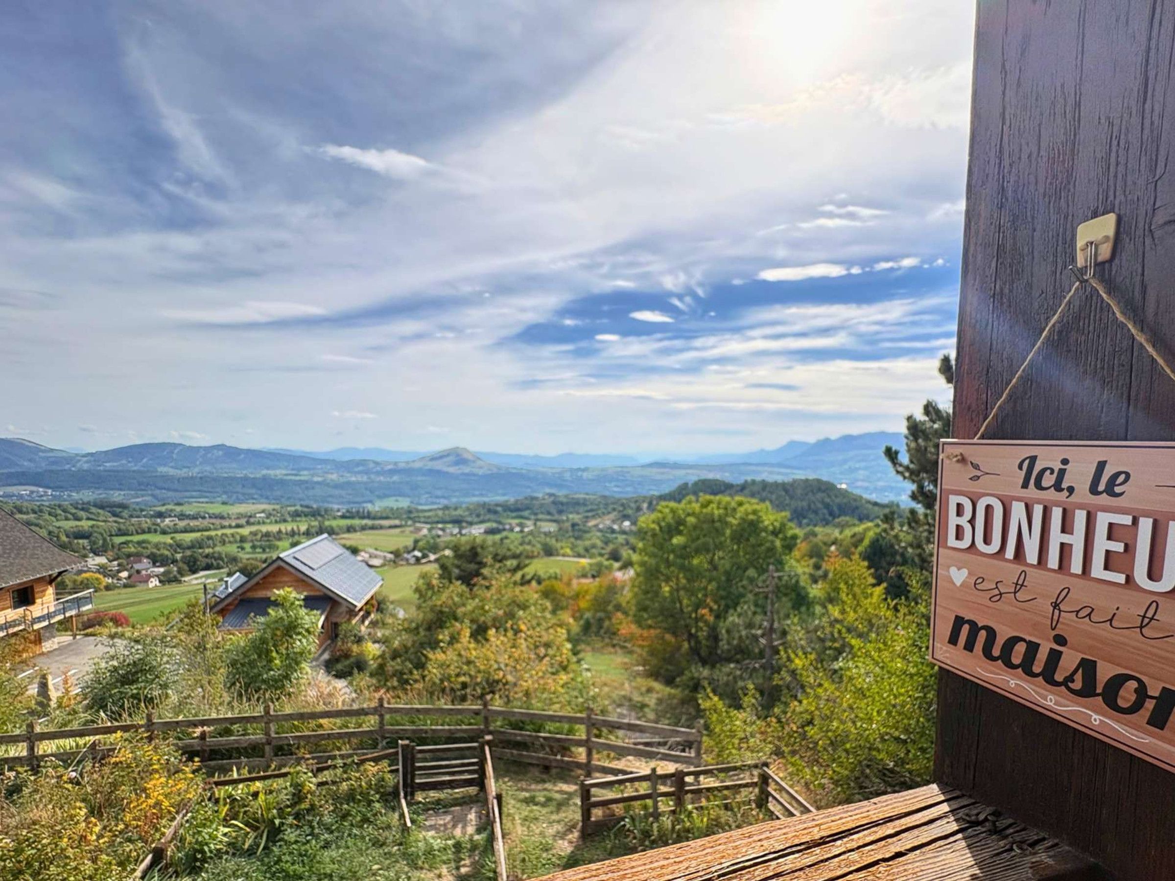 A wooden sign with text, a wooden fence, and a view of a house and landscape.