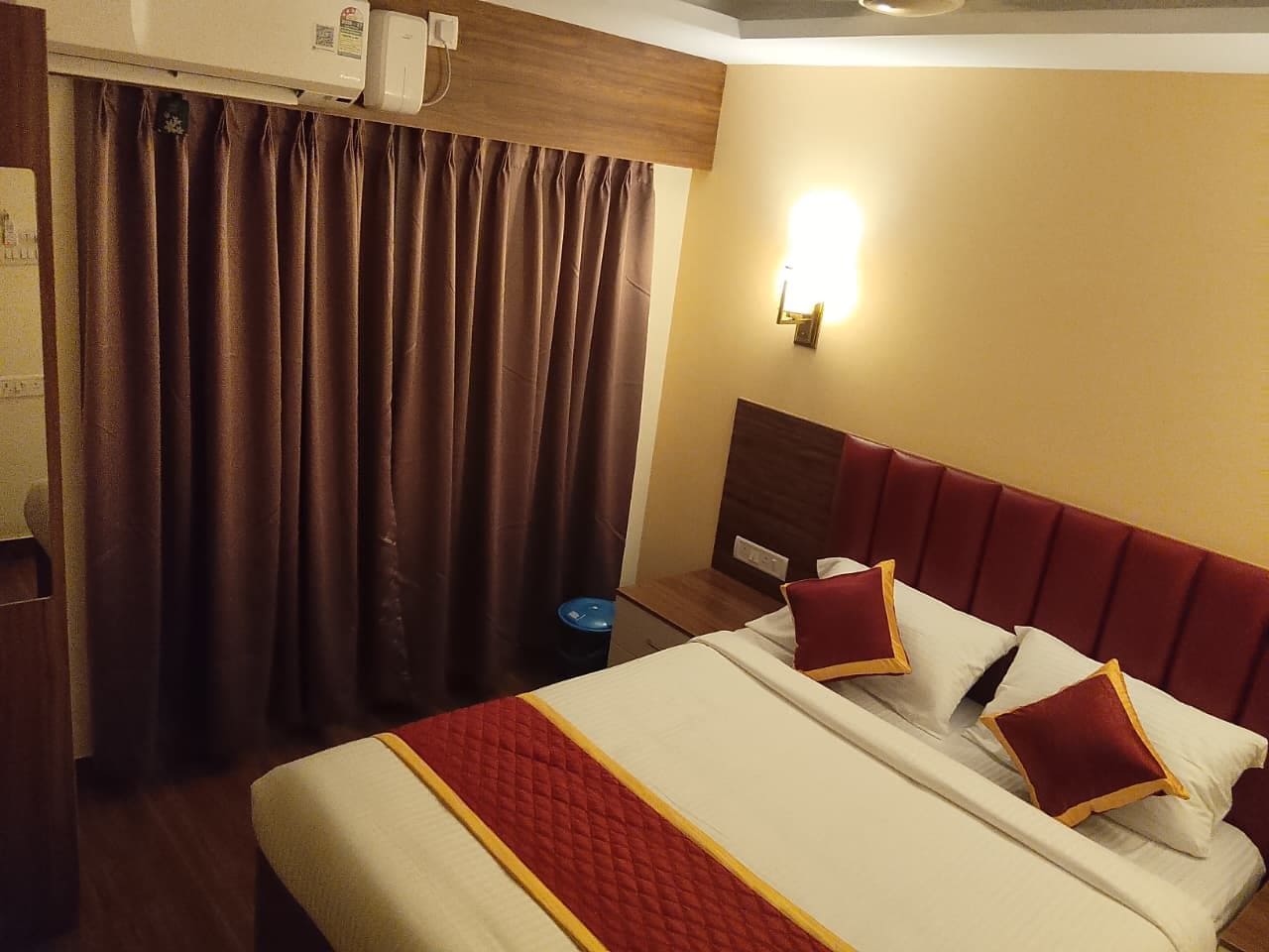 Una habitación de hotel con cama, cortinas, una lámpara fijada a la pared y un cubo de basura pequeño.