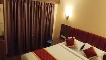 Una habitación de hotel con cama, cortinas, una lámpara fijada a la pared y un cubo de basura pequeño.