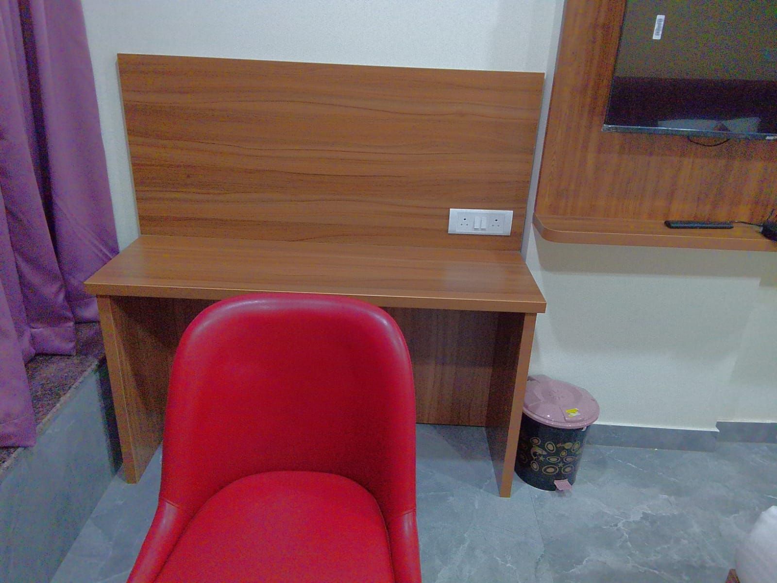 Una habitación con un escritorio de madera, una silla roja, un cubo de basura y una ventana.