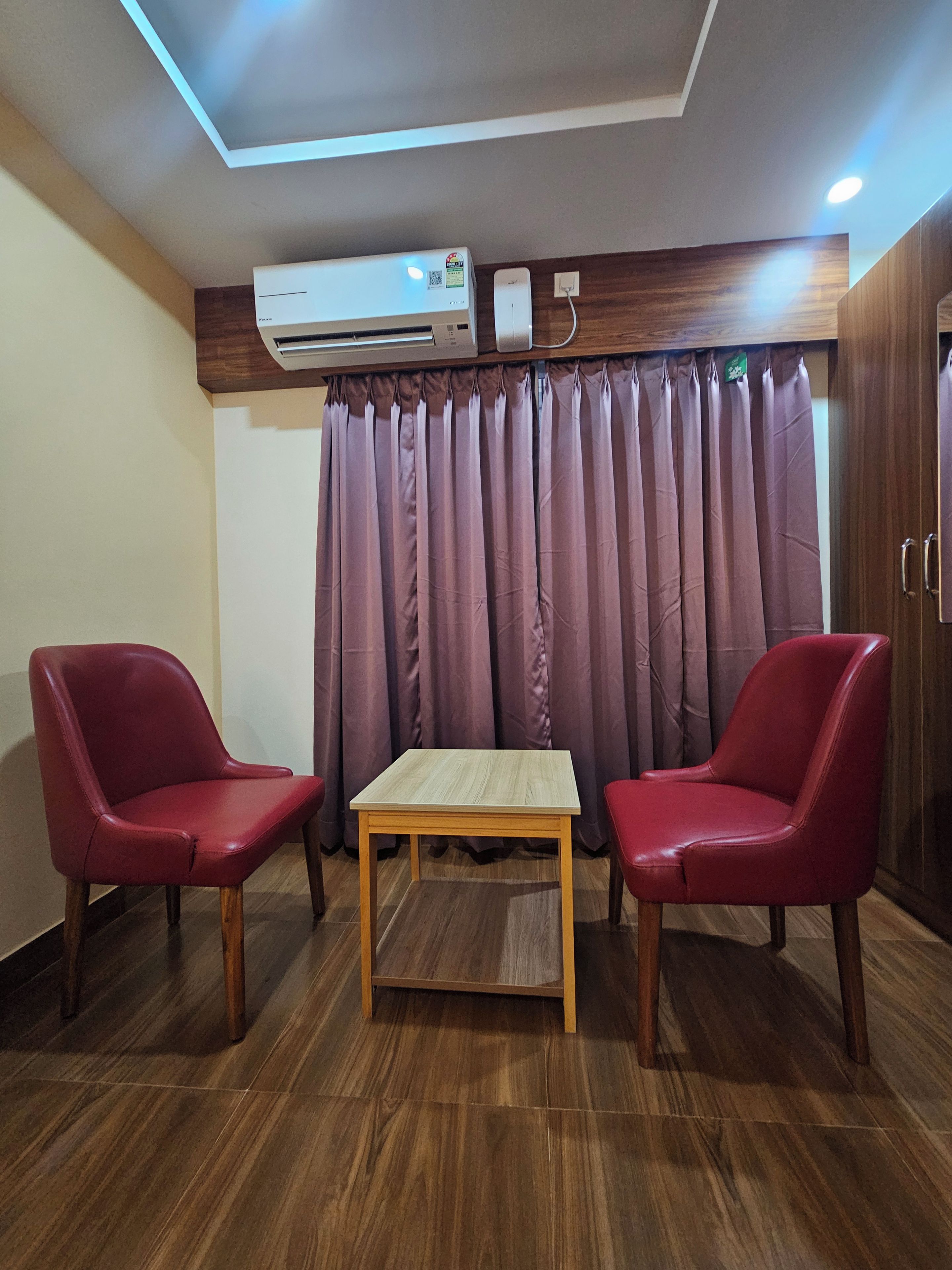 Una habitación con piso de madera, dos sillas rojas, una mesita de madera, un equipo de aire acondicionado y una cortina.