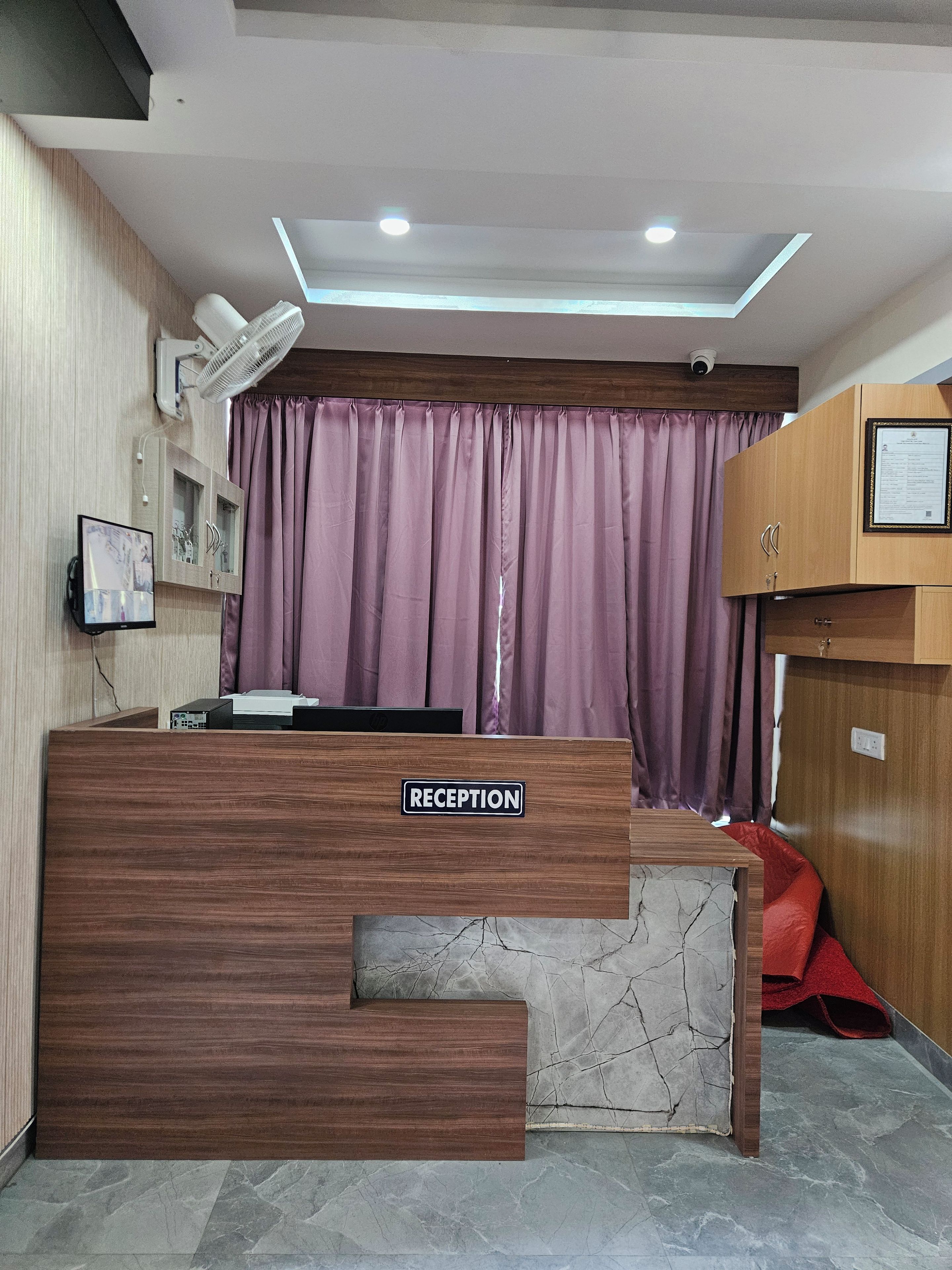 A reception desk with a 'RECEPTION' sign, a mounted fan, a security camera, and a wooden cabinet with a framed certificate.