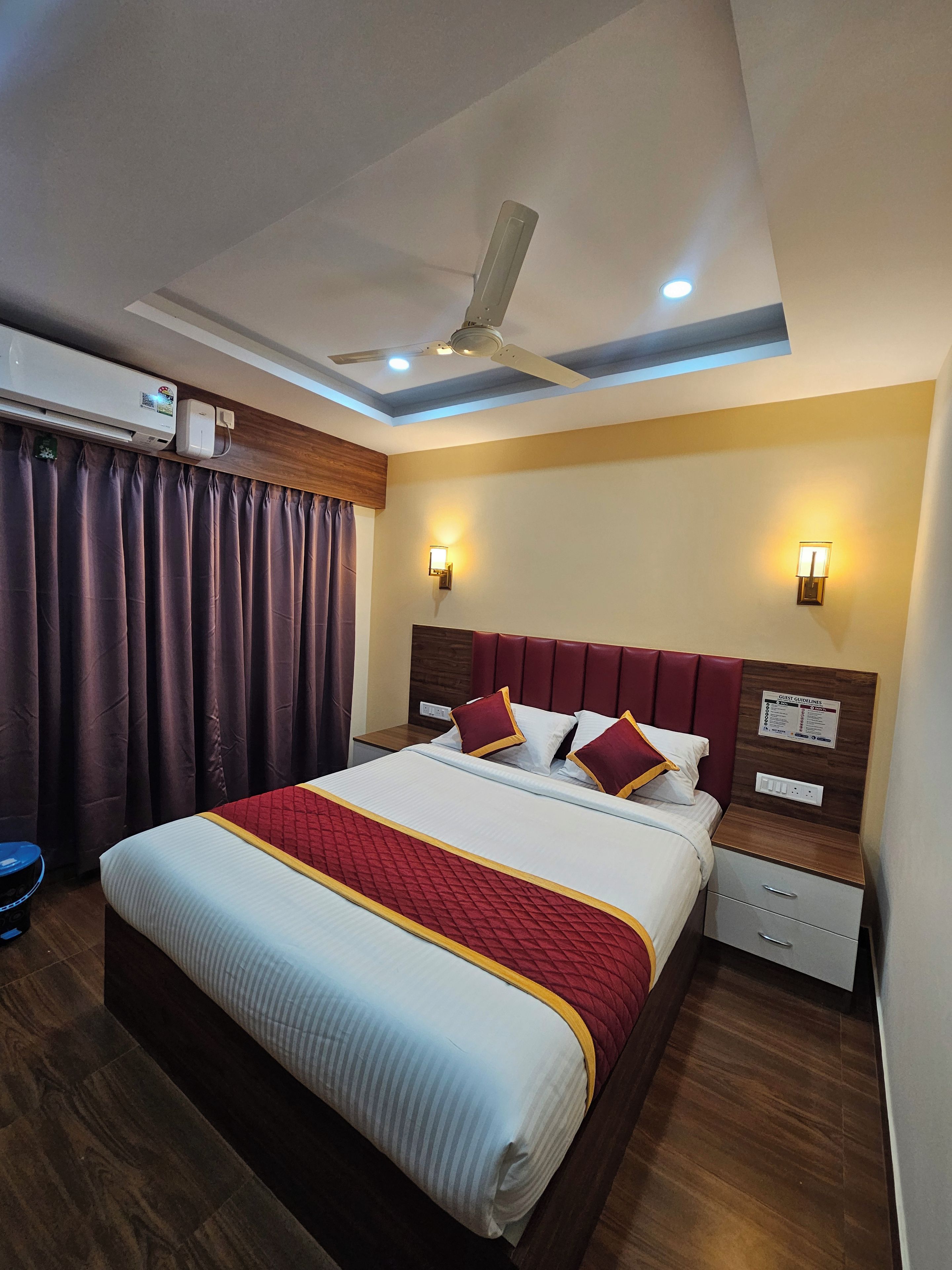Habitación de hotel con cama, ventilador de techo, equipo de aire acondicionado, mesita de noche, lámpara de pared y un cartel con normas para huéspedes.