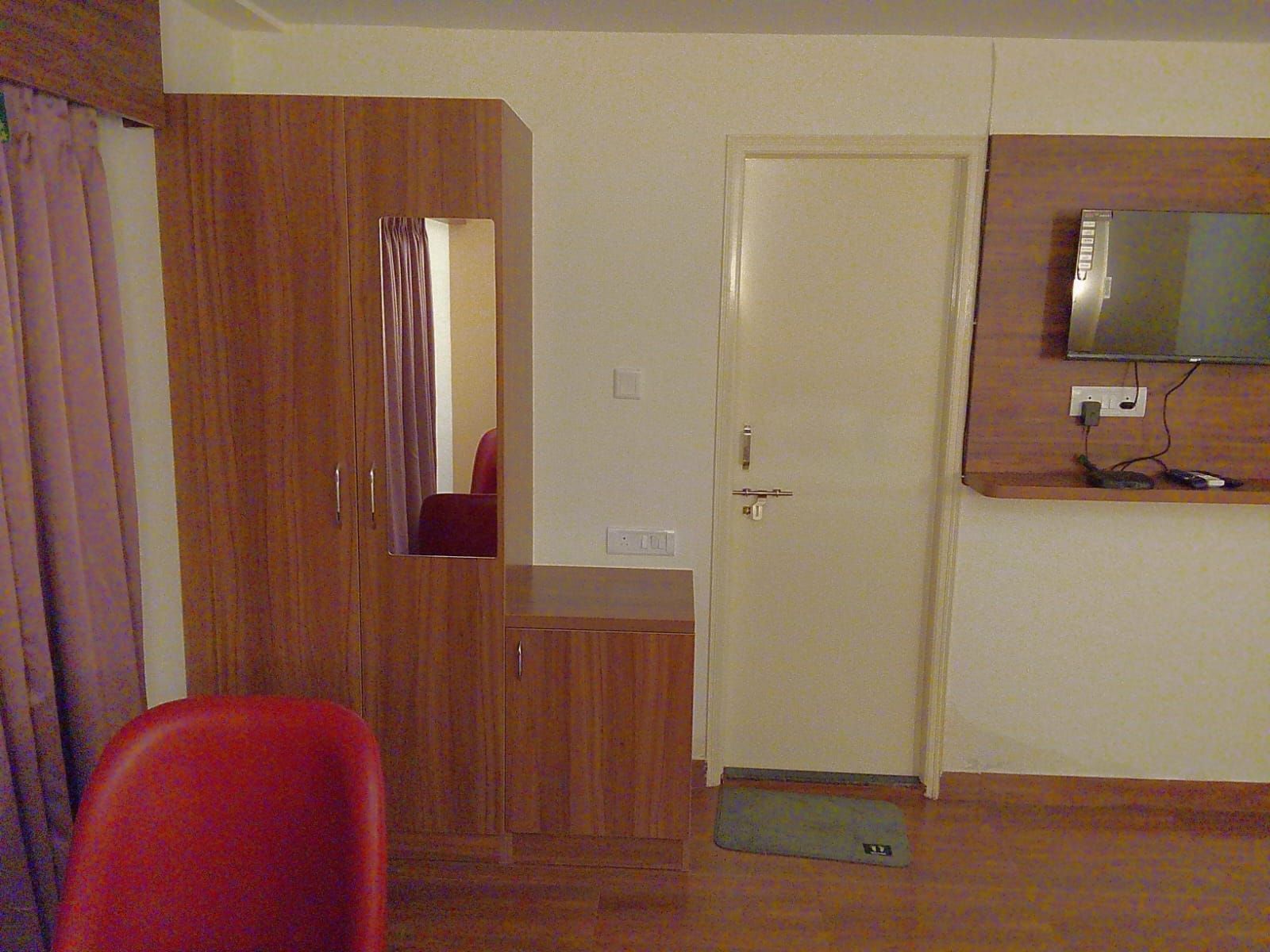 Una habitación con un armario de madera, una silla roja, un televisor montado en la pared y una puerta con un felpudo en el suelo.