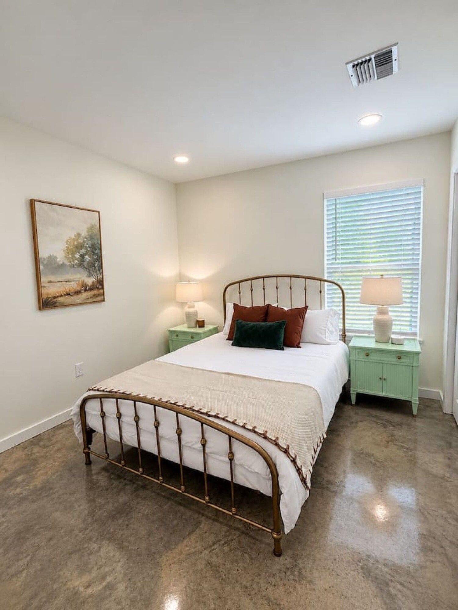 A bedroom with a metal-framed bed, a nightstand with a lamp, a window with blinds, and a painting on the wall.