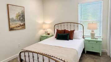 A bedroom with a metal-framed bed, a nightstand with a lamp, a window with blinds, and a painting on the wall.